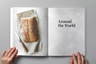 Photo of a designer holding the cookbook open, showing a section divider with a picture of a bread and the heading ‘Around the World’.
