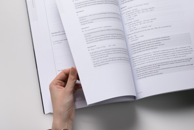 Photograph with a hand holding open a Practice Exams book showing answers to questions.