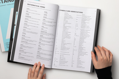 Photograph showing hands holding open a student book to reveal a double-page spread showing mathematical verbs and symbols.